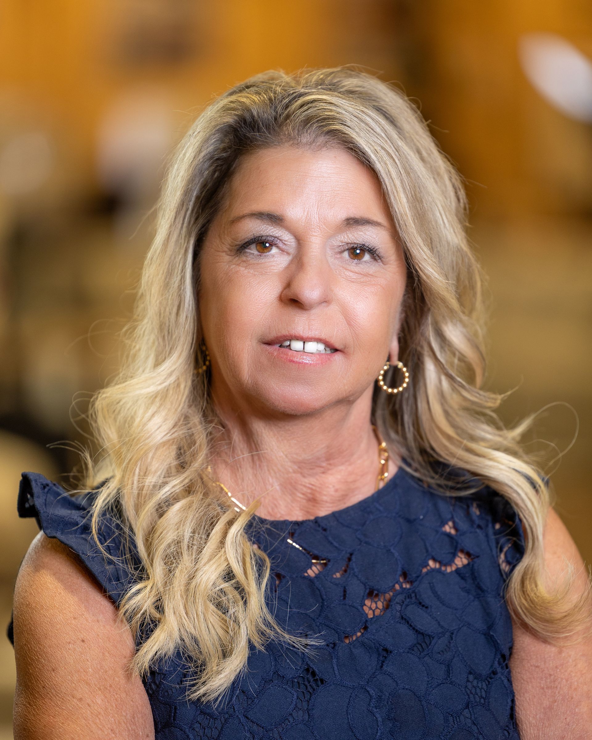 Woman with blonde wavy hair, wearing a blue lace top, smiling, gold earrings, indoors.