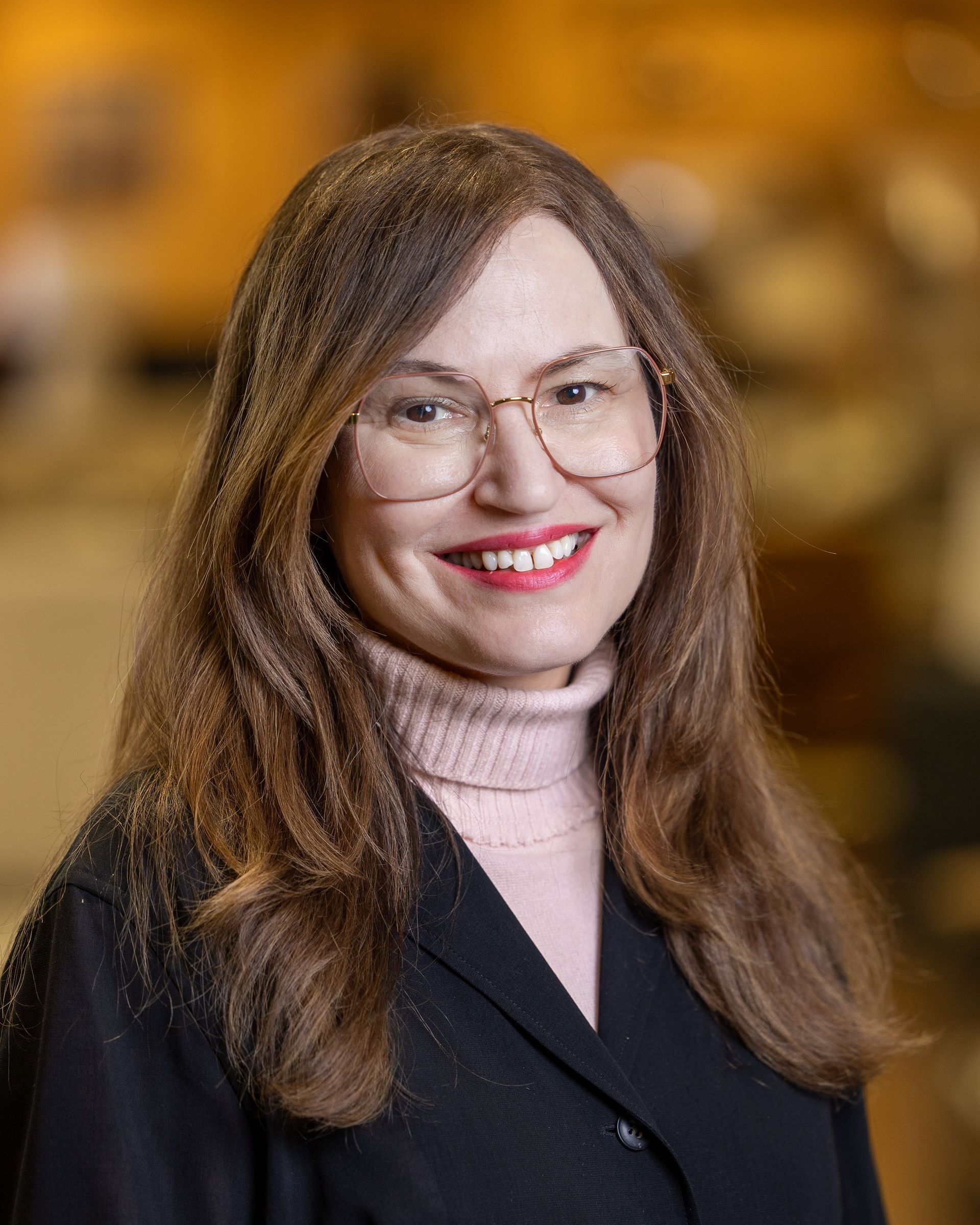 Woman with long brown hair smiles, wearing glasses, pink turtleneck, and black blazer.