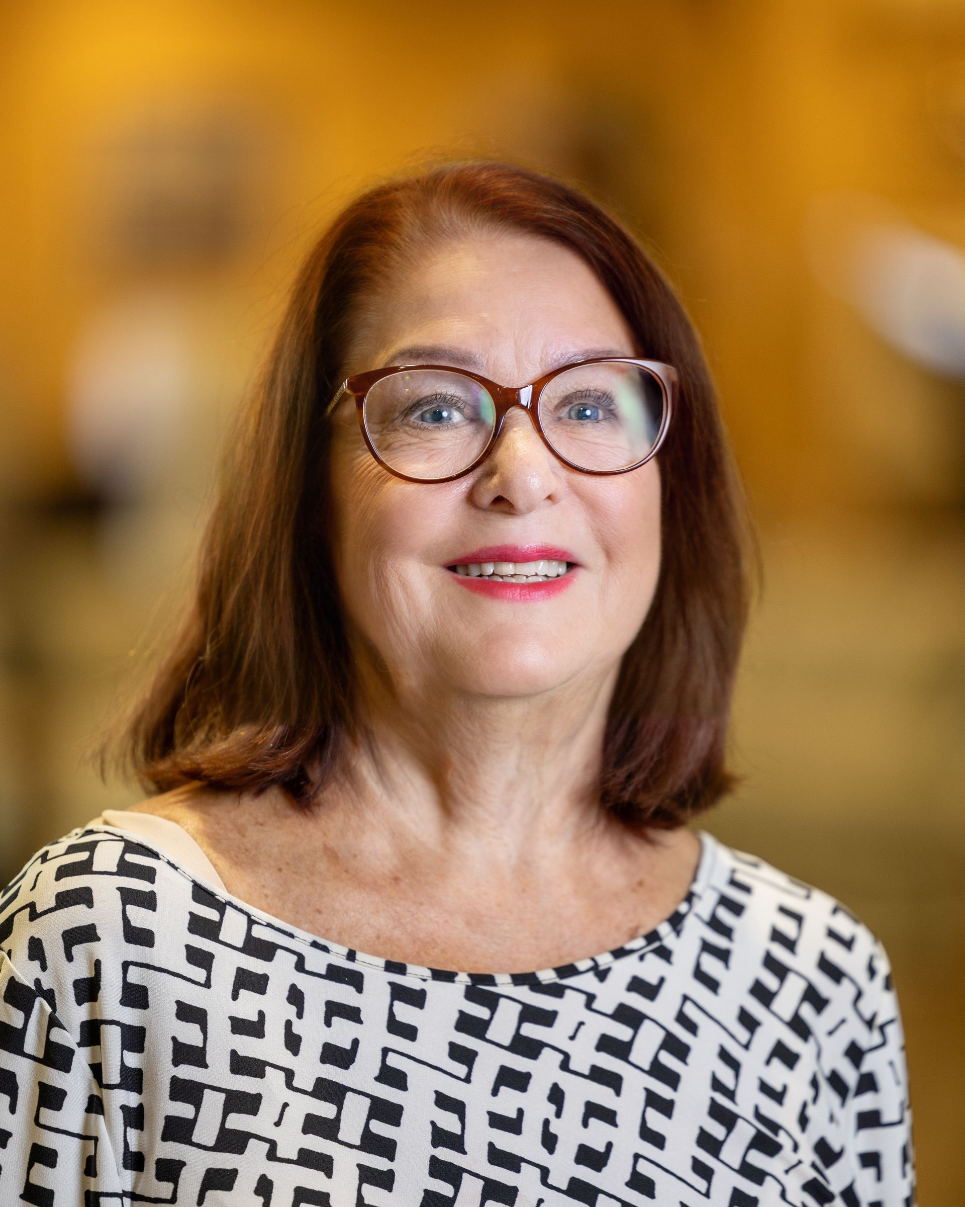 Woman with glasses, smiling, wearing a black and white patterned top, brown hair. Soft, yellow background.