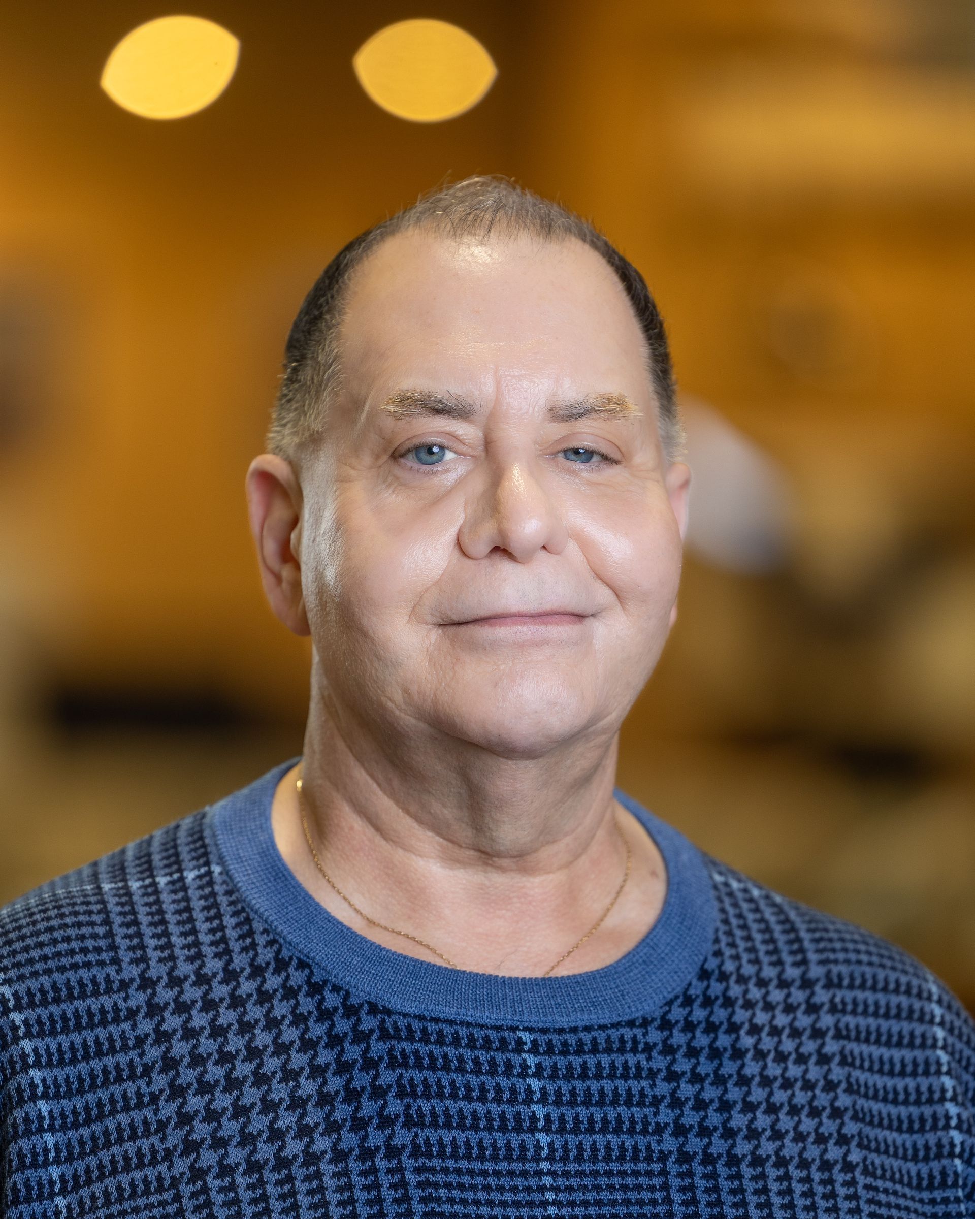 Man in blue sweater smiles, gold necklace, neutral background, indoors.