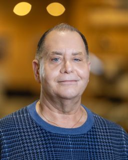 Man in blue sweater smiles, gold necklace, neutral background, indoors.