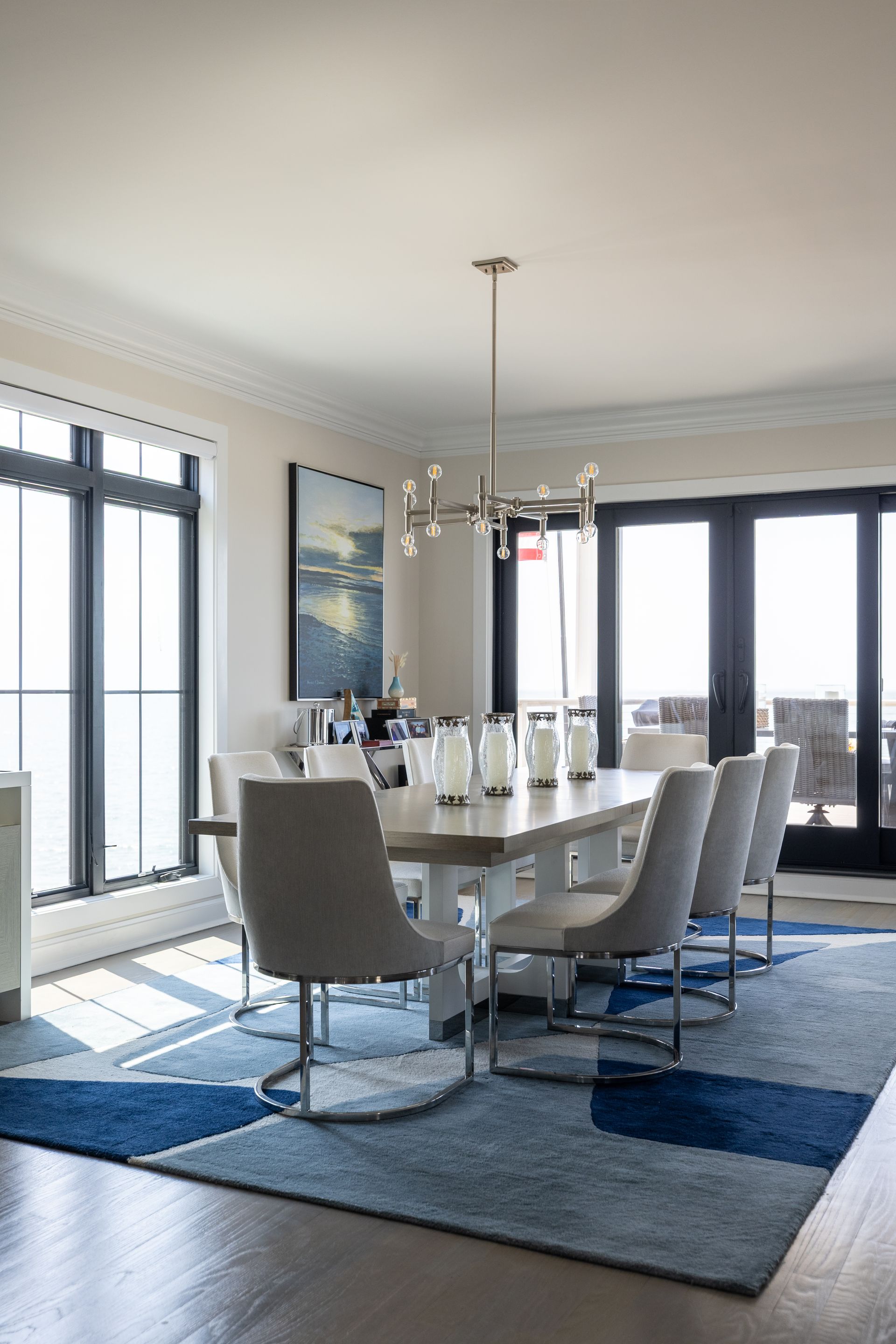 Elegant dining room with rectangular table, upholstered chairs, blue geometric rug, and large windows.