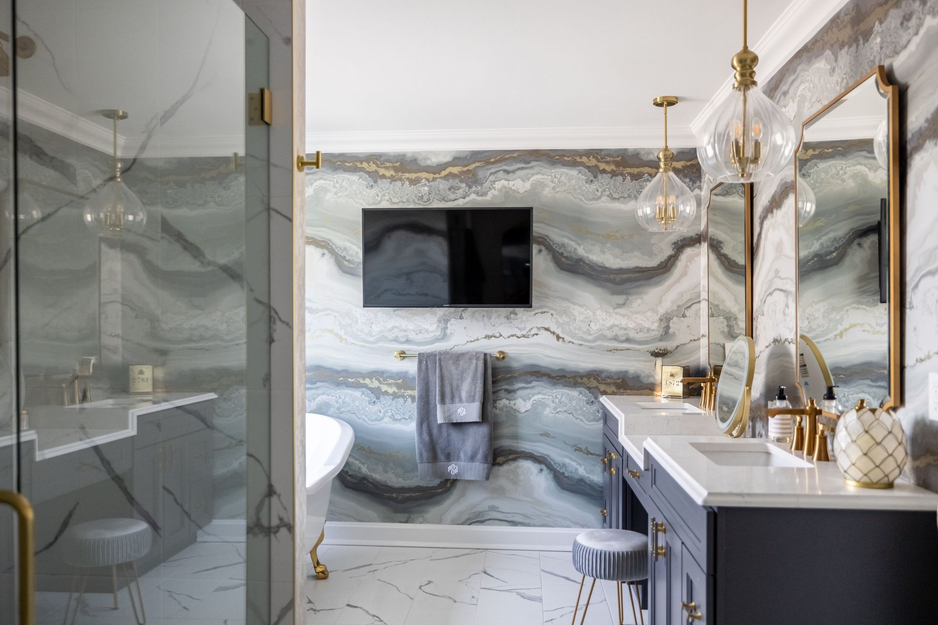 Luxurious bathroom with marble-effect wall, dark vanity, and gold accents. Includes TV, mirrors, and glass shower.