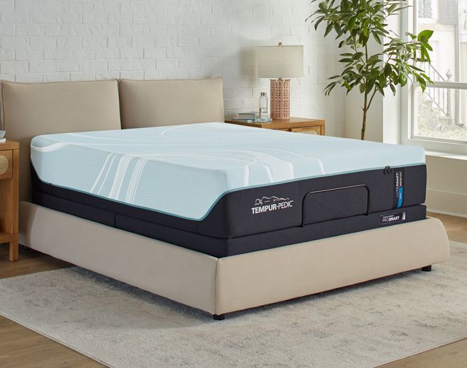 Woman with blonde hair smiles at the camera, wearing a black top and silver necklace. Blurred indoor setting.