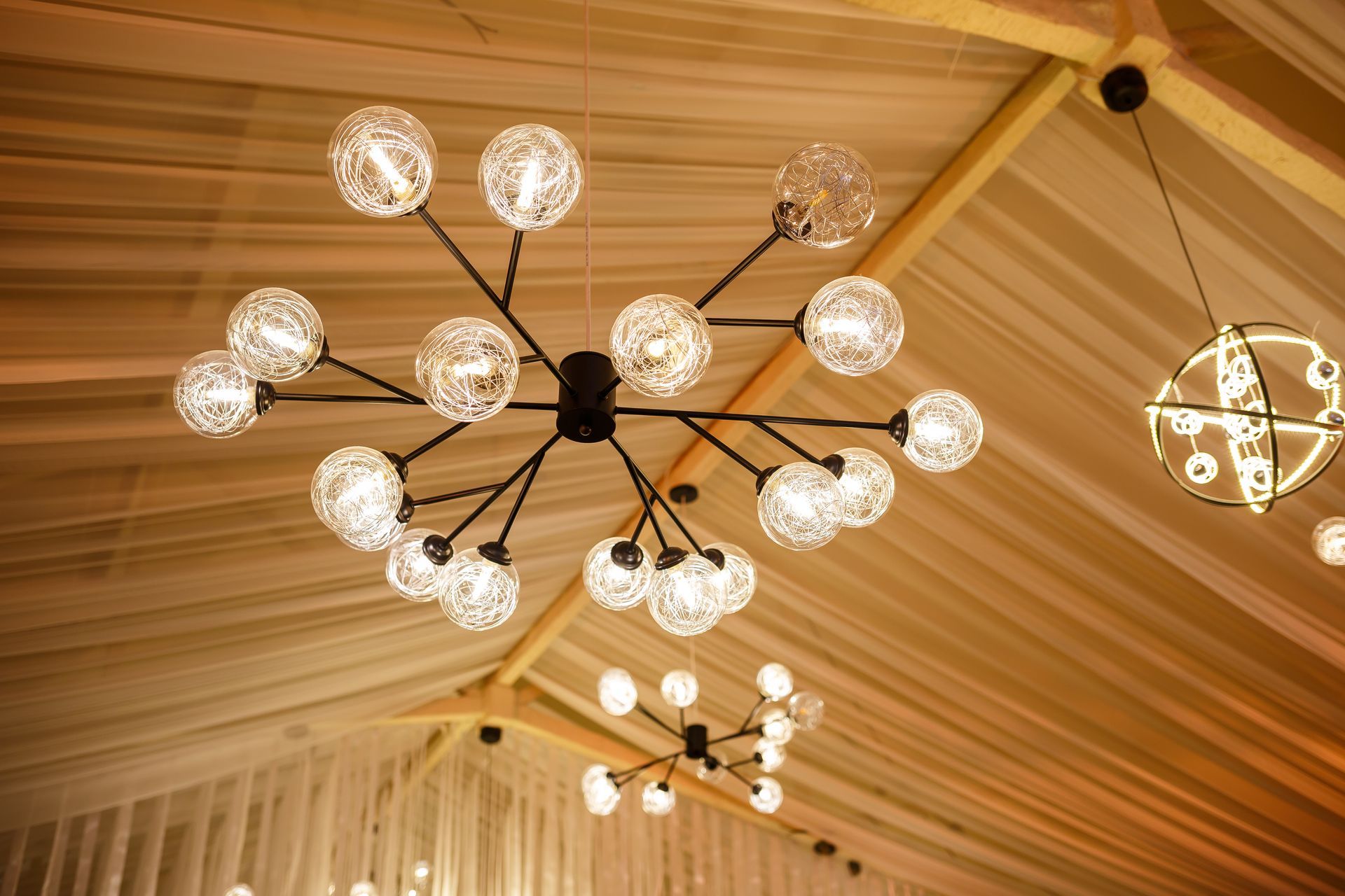 Black chandelier with multiple globe lights hangs from a tent ceiling with soft, warm lighting.