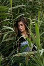 Woman in a black shirt and denim overalls surrounded by tall green plants, looking down.