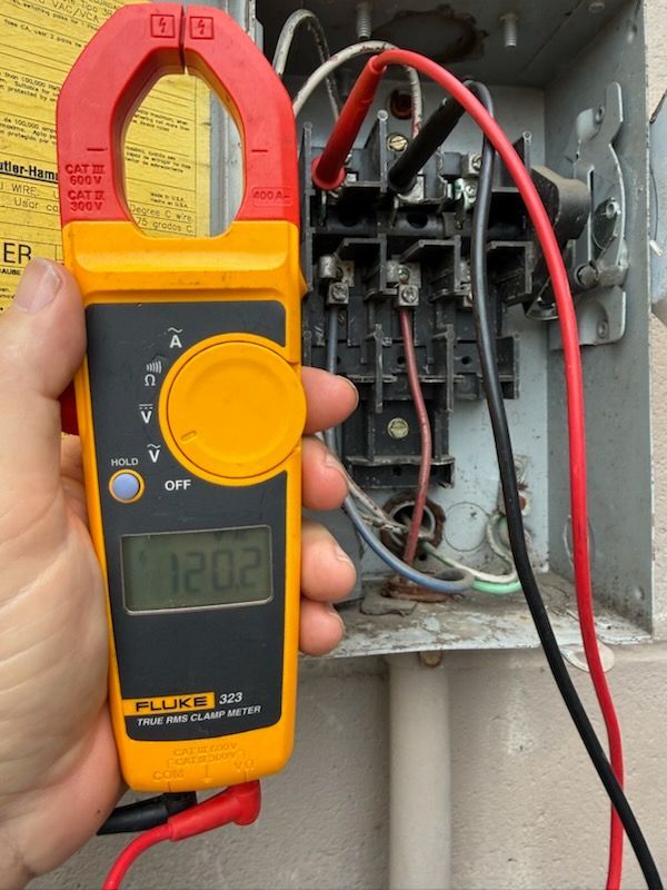 A hand holds a yellow Fluke multimeter, showing a 120.2V reading while probing terminals inside an open electrical box.