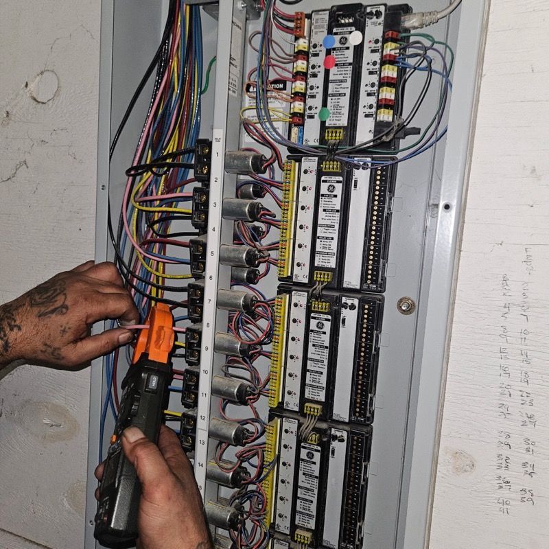 A person uses a digital clamp meter to test electrical wiring inside an industrial lighting control panel.
