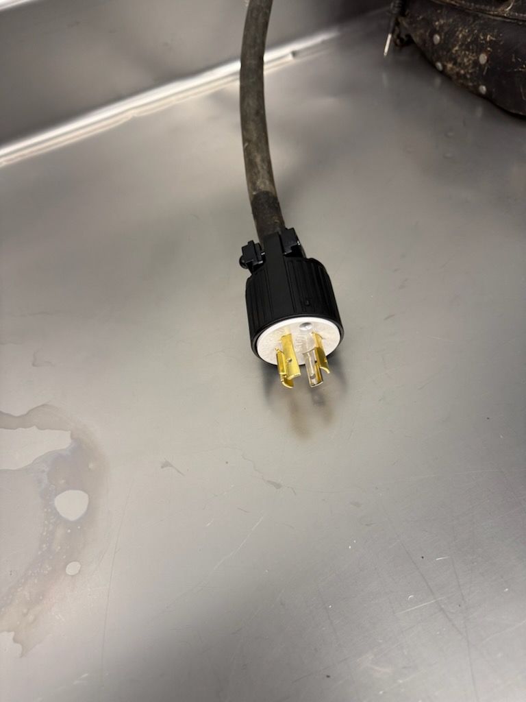 A black industrial-style NEMA power plug with three metal prongs, resting on a flat, gray metallic surface.