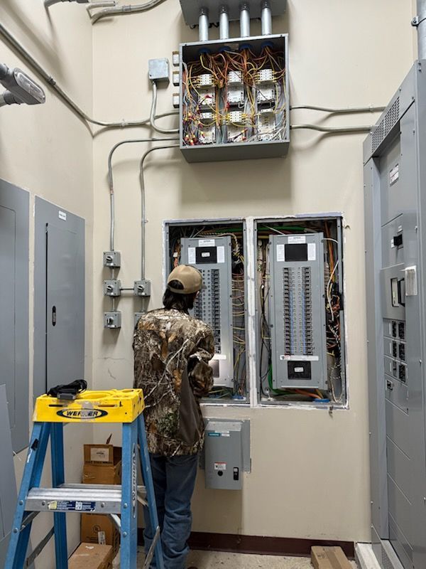 A person in camouflage clothing stands on a ladder working on an open electrical panel in a room with industrial equipment.
