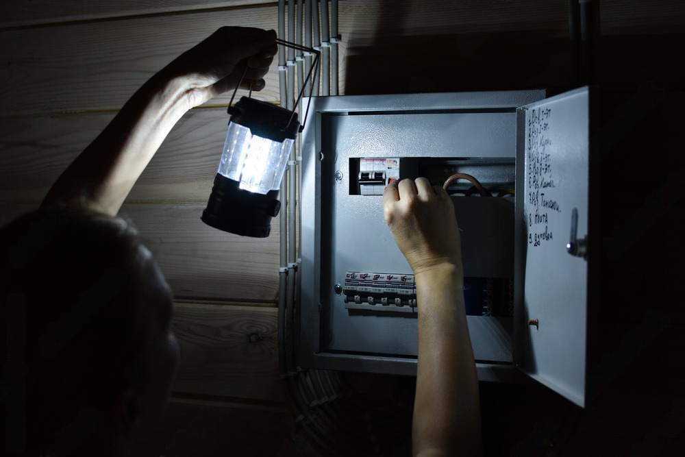 A person uses a handheld lantern to illuminate an open electrical breaker box in a dimly lit, wood-walled room.