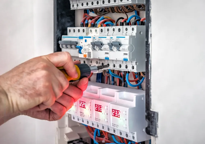 A hand uses a yellow and black screwdriver to adjust circuit breakers in an electrical panel.