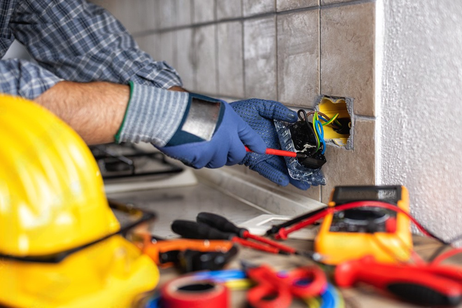 An electrician in blue gloves uses a screwdriver to work on exposed electrical wiring in a tiled wall.