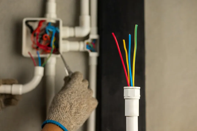 A gloved hand uses a tool to connect multicolored wires inside a junction box mounted on a gray wall with plastic conduit.