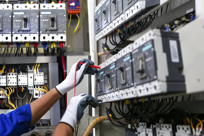 An electrician in work gloves uses a multimeter probe to test a circuit breaker panel.
