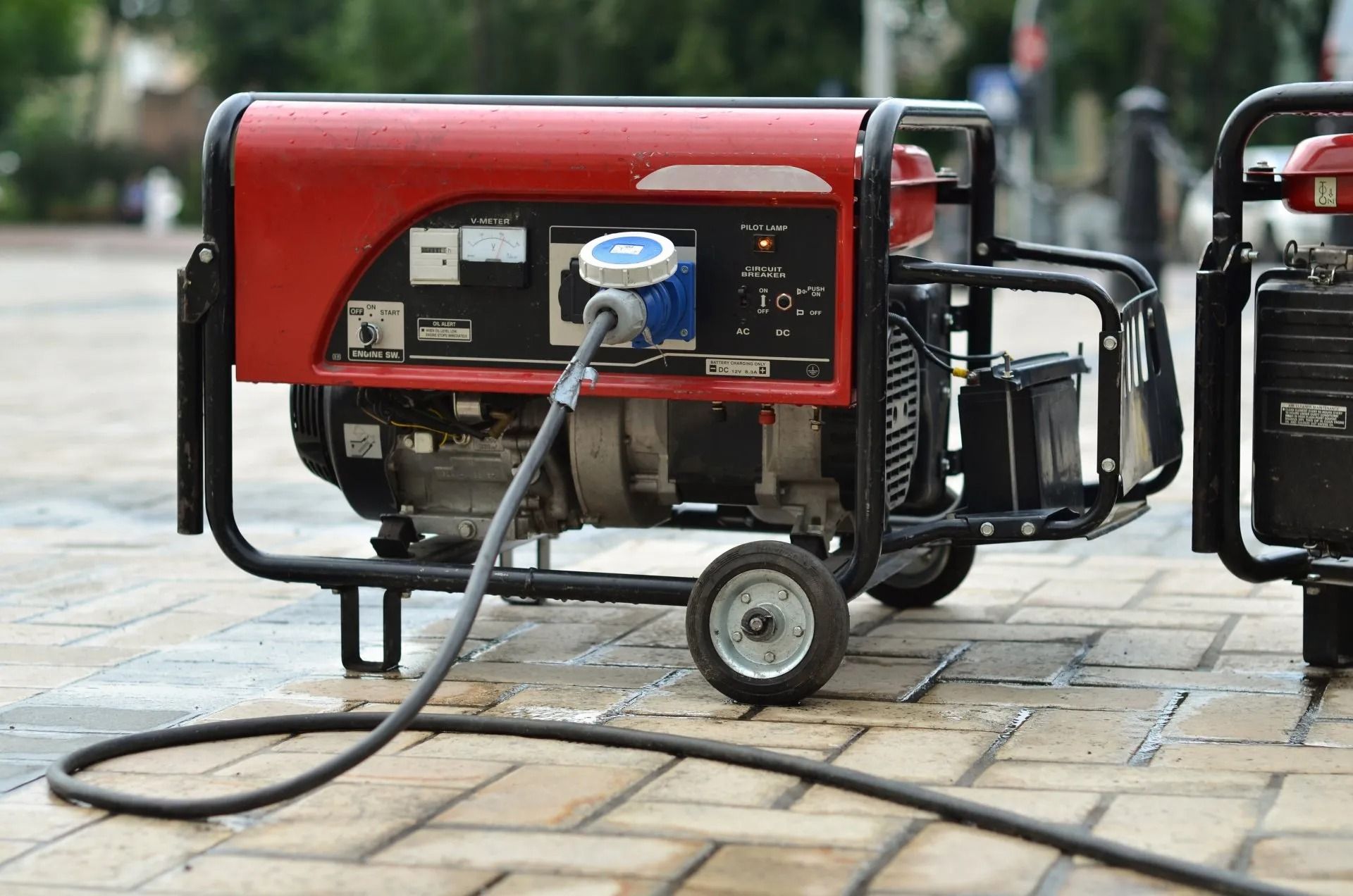 A red portable power generator sits on a paved outdoor surface, with a power cord plugged into its front panel.