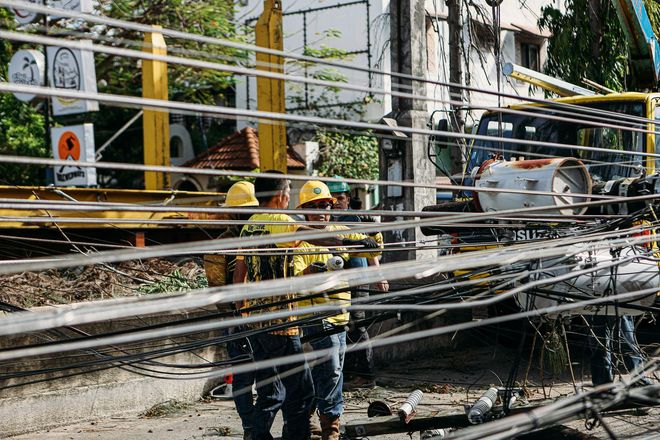 Utility workers in hard hats and yellow high-visibility vests work on overhead power lines near a street.