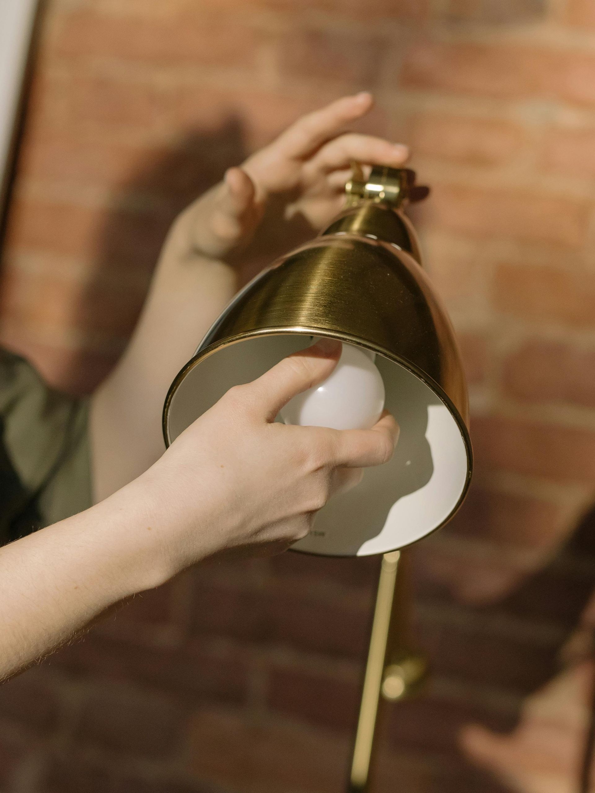 Hands adjusting a white light bulb inside a gold metal lamp shade against a brick wall background.