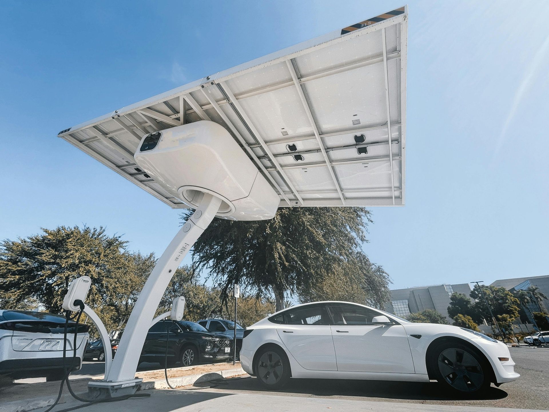 A white electric car parked under a modern, tilted solar panel charging station on a sunny day.