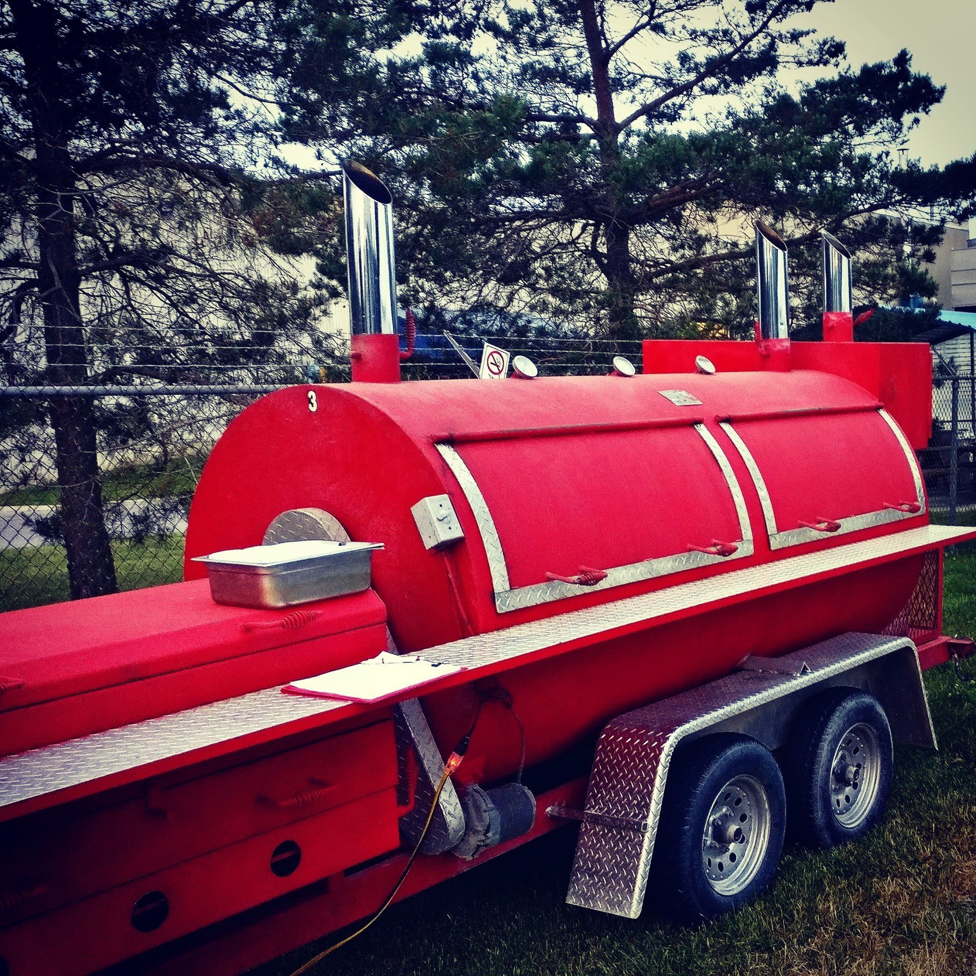 A large, bright red BBQ smoker mounted on a trailer, equipped with multiple smoke pipes and metallic latches