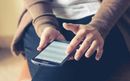 Person using a smartphone; dark screen, touching the screen with one finger. Wears a brown cardigan and dark pants.