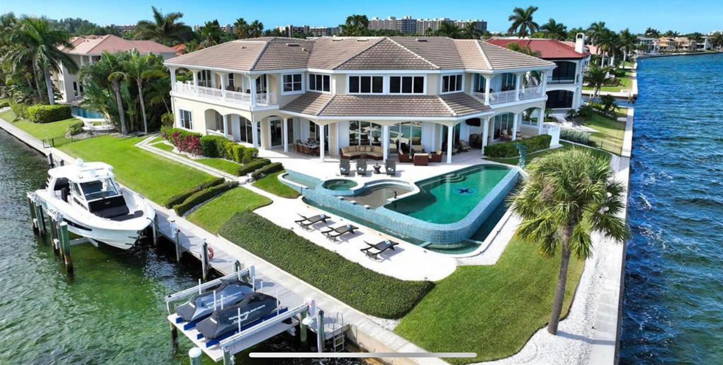 Luxury waterfront home with pool, dock, and boats. White exterior, blue water and sky.