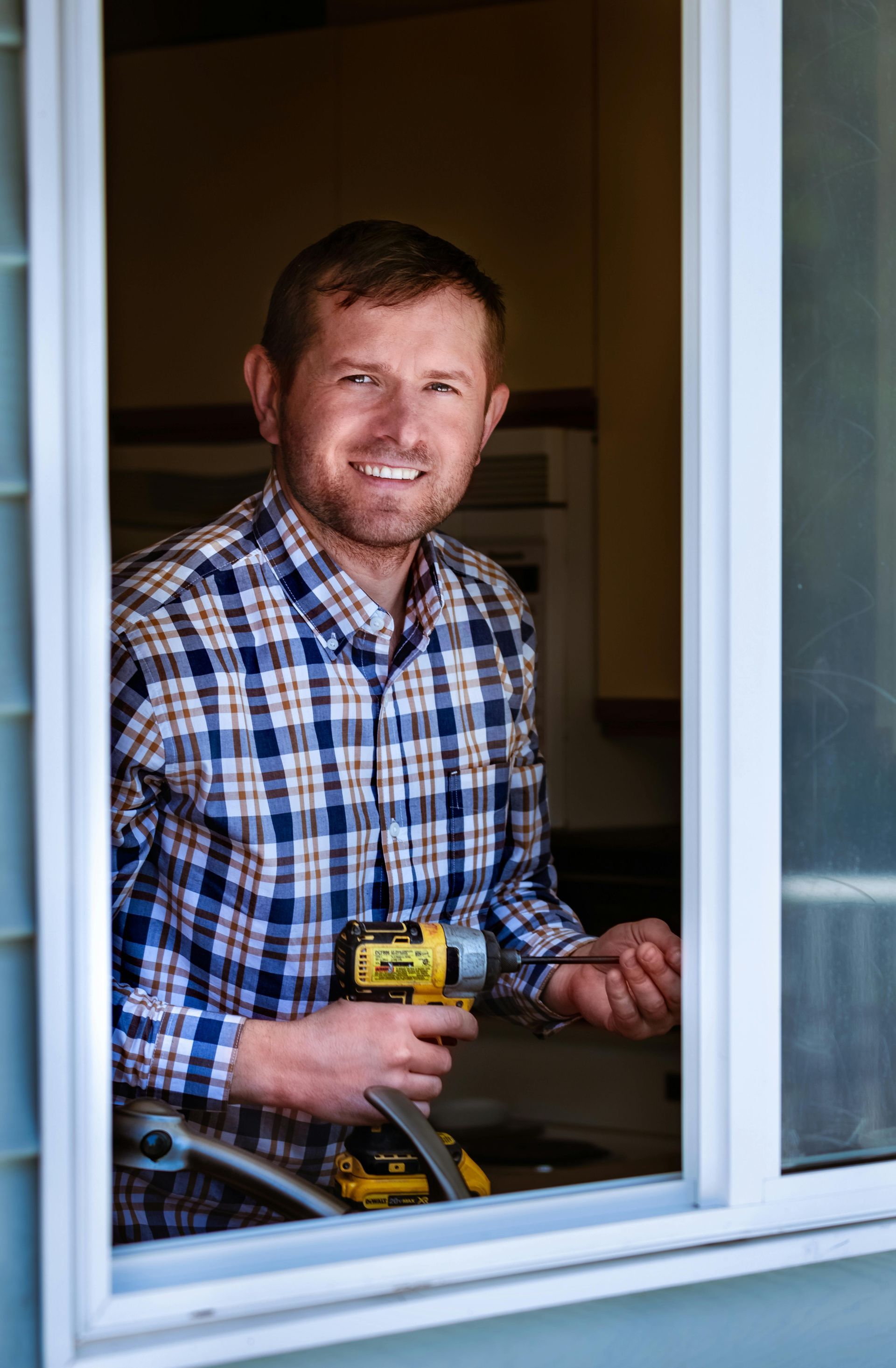 Man holding a drill smiles, framed by a window. He wears a plaid shirt, standing in a kitchen with blue siding.