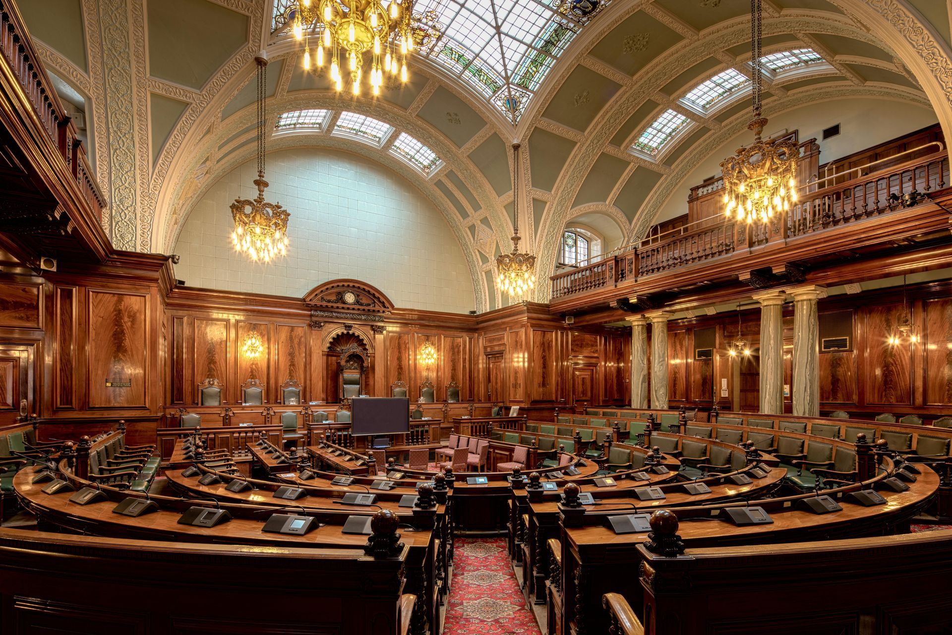 Ornate parliamentary chamber with rows of desks, wood paneling, chandeliers, and a vaulted ceiling