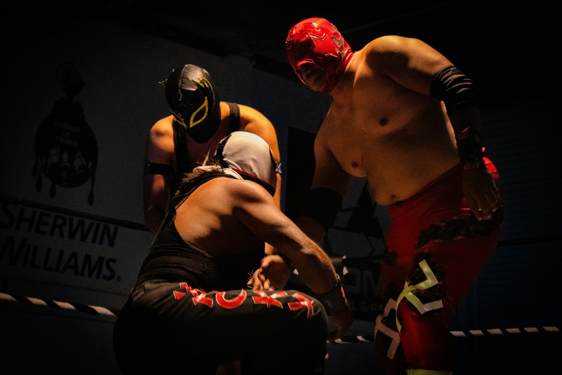 Three masked wrestlers in a wrestling ring, two are confronting a third. The red masked wrestler is shirtless.