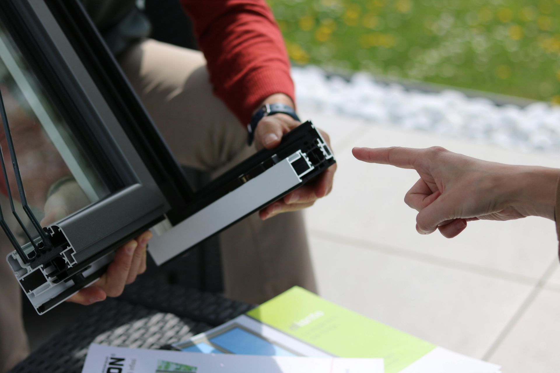 Person holding a window frame, another pointing. Outdoors, product demonstration.