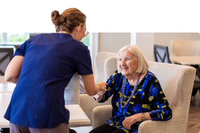 Caregiver assisting an elderly person sitting in a chair. Both are smiling.