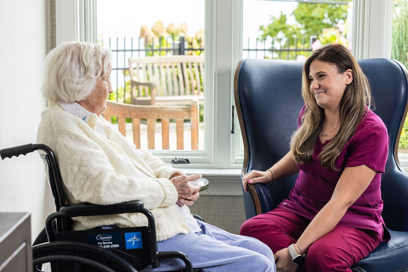 Two women chatting in a bright room, one seated in a wheelchair by a window.