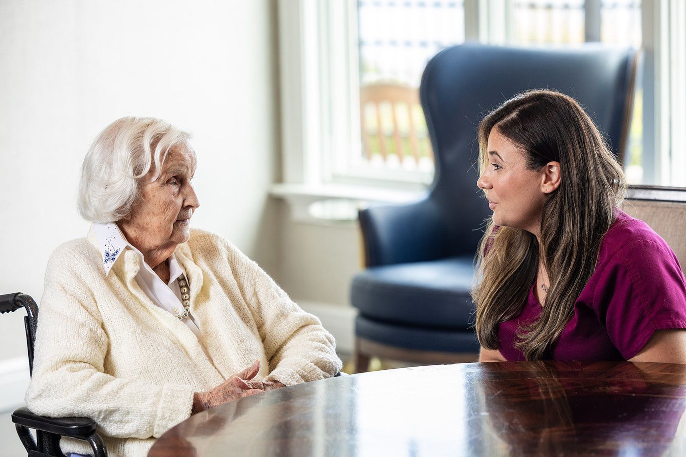 Two women talking at a table in a bright office, with a blue armchair in the background