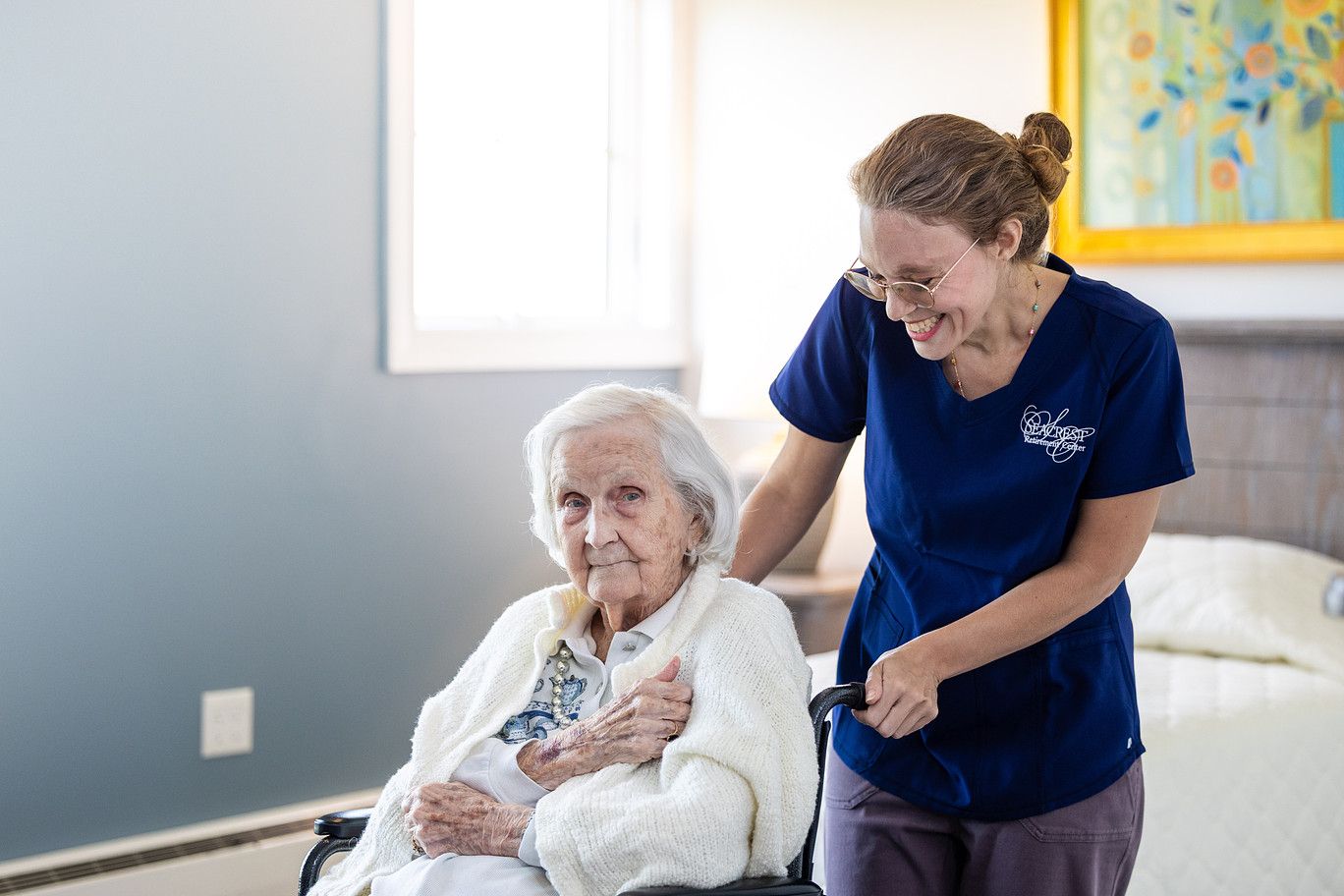 Nurse in blue scrubs assisting an elderly patient seated in a bright bedroom