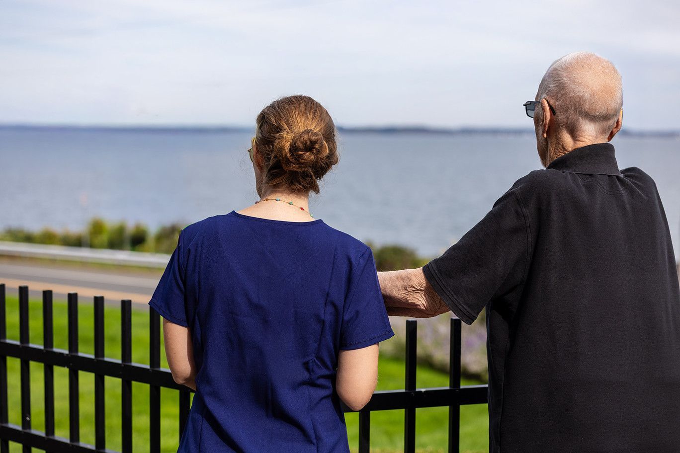 Two people stand by a black fence overlooking a calm body of water on a sunny day.