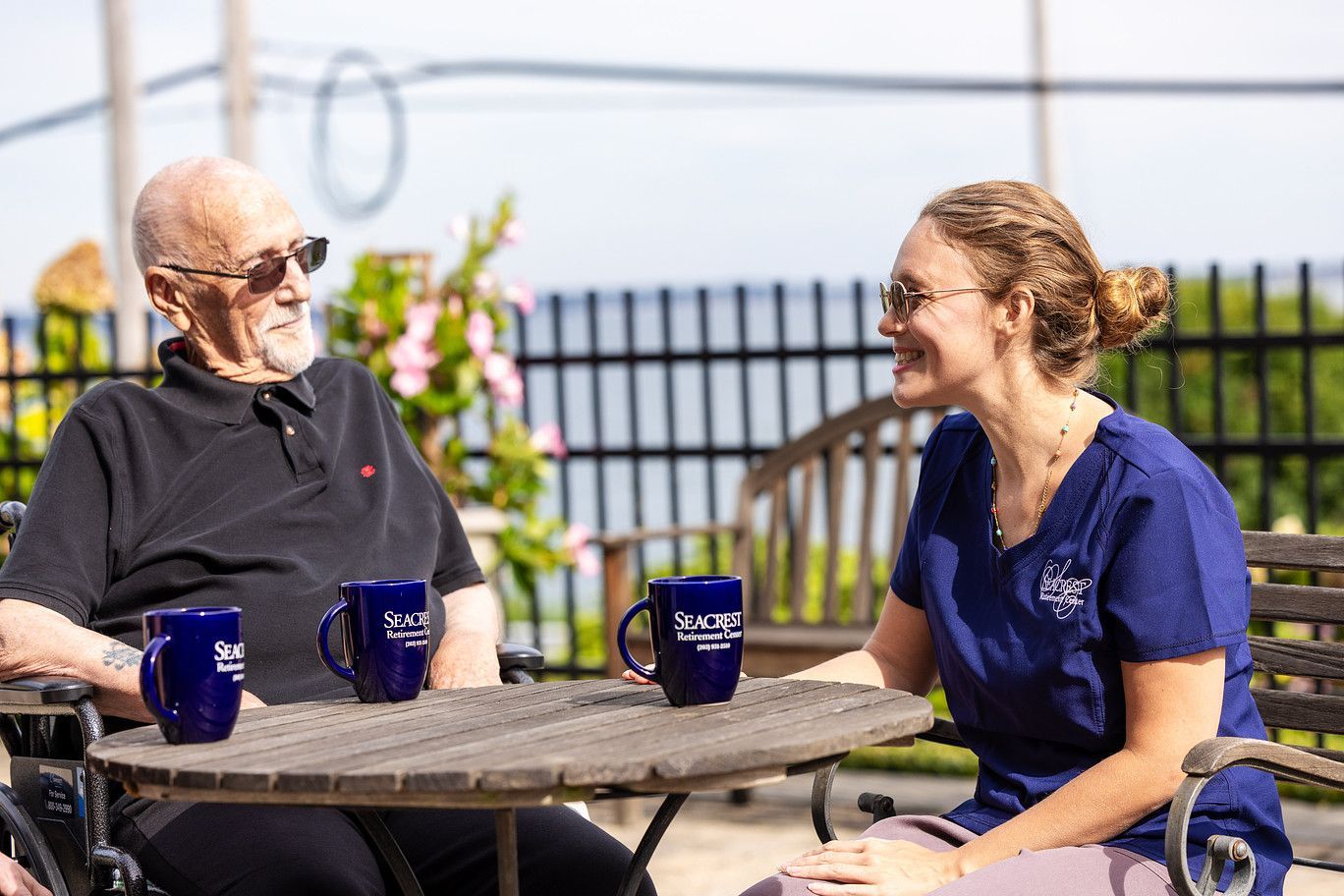 Two people chat at an outdoor table with blue mugs, smiling in a sunny patio setting.