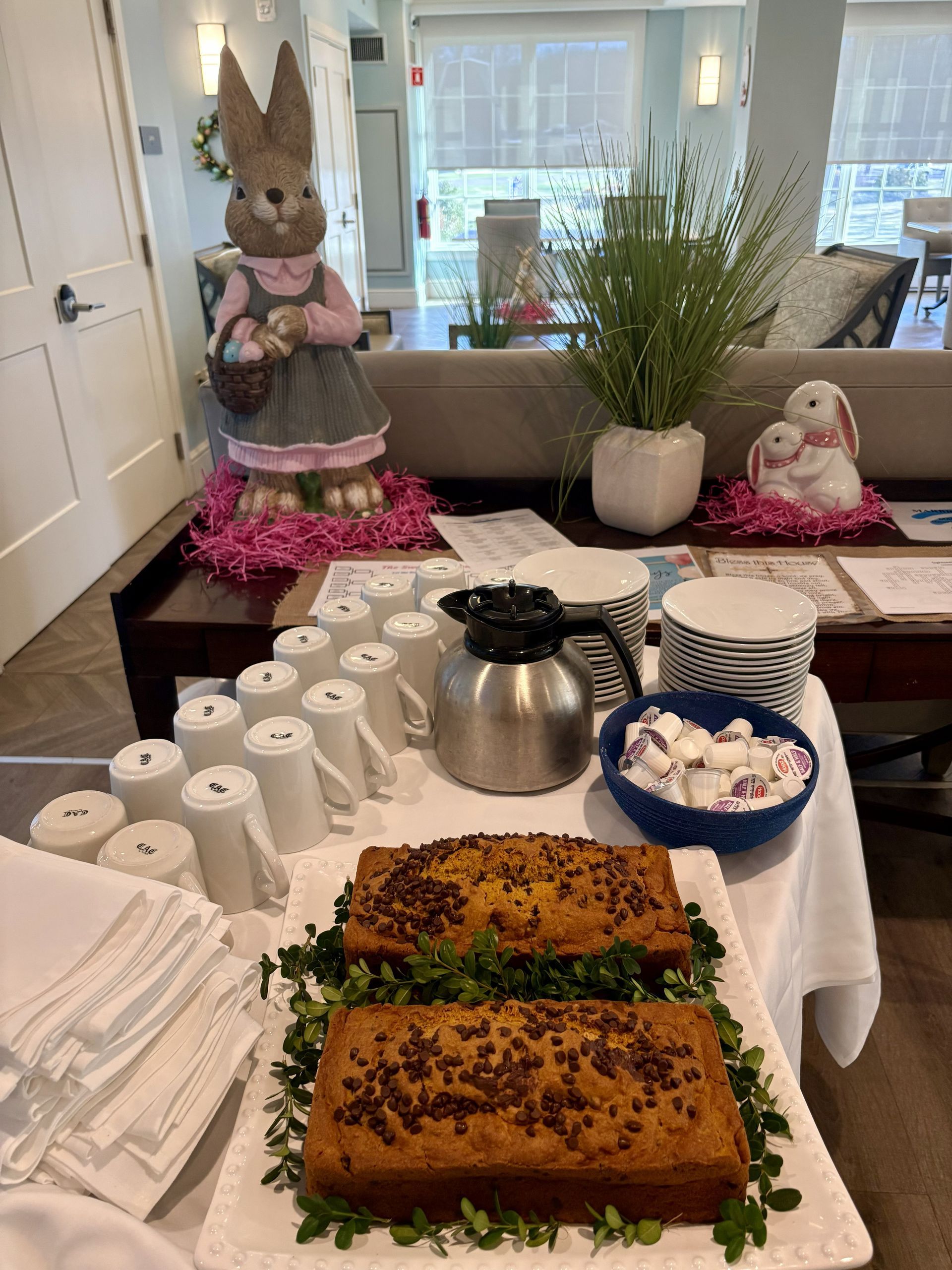 Easter-themed buffet featuring loaves of bread, coffee, mugs, and bunny decorations on a white-clothed table.