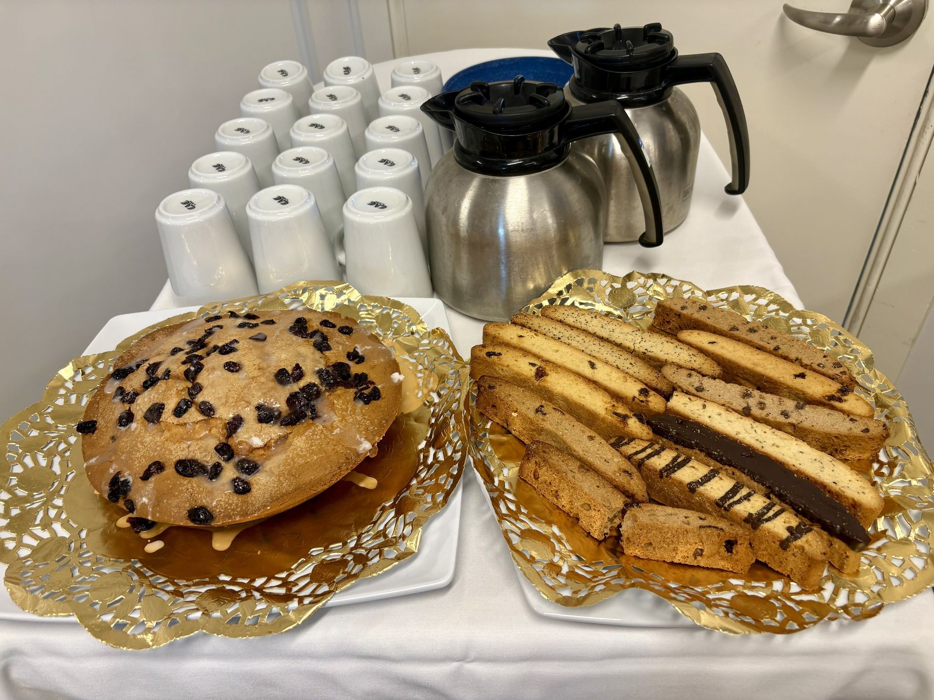 Dessert trays with a chocolate chip pastry and sliced cake beside coffee pots and stacked cups
