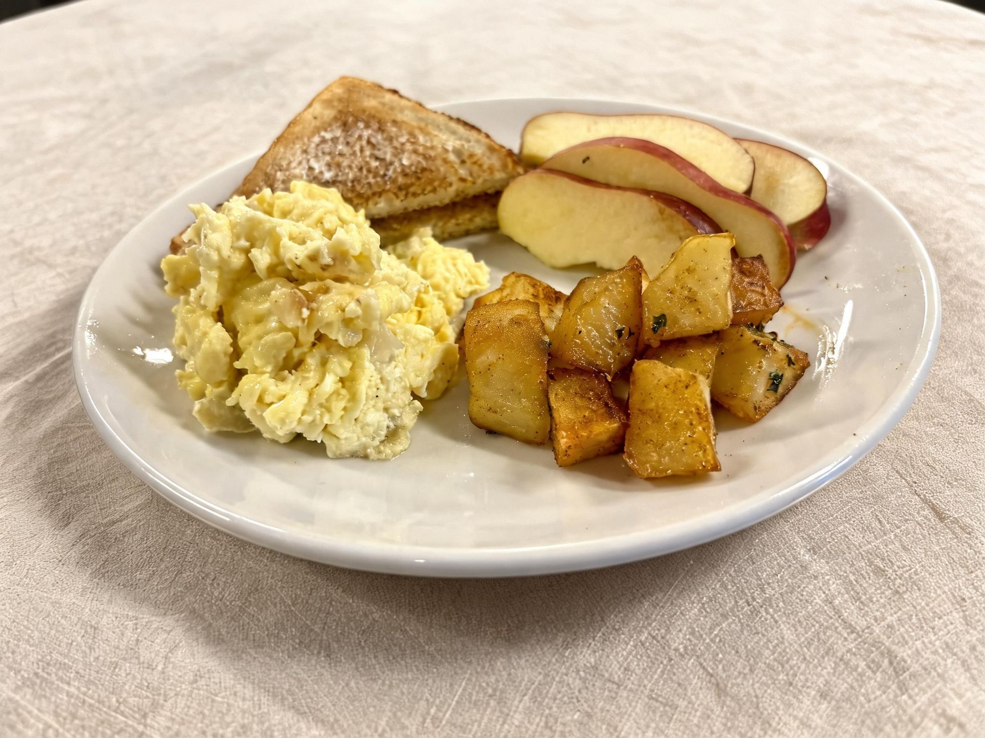 Plate with scrambled eggs, toast, apple slices, and roasted potato wedges on a table