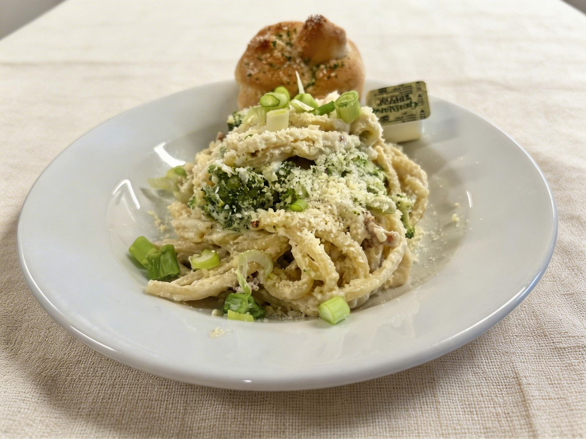 A white bowl of creamy pasta topped with grated cheese and chopped green onions, served with a small bread roll and butter.