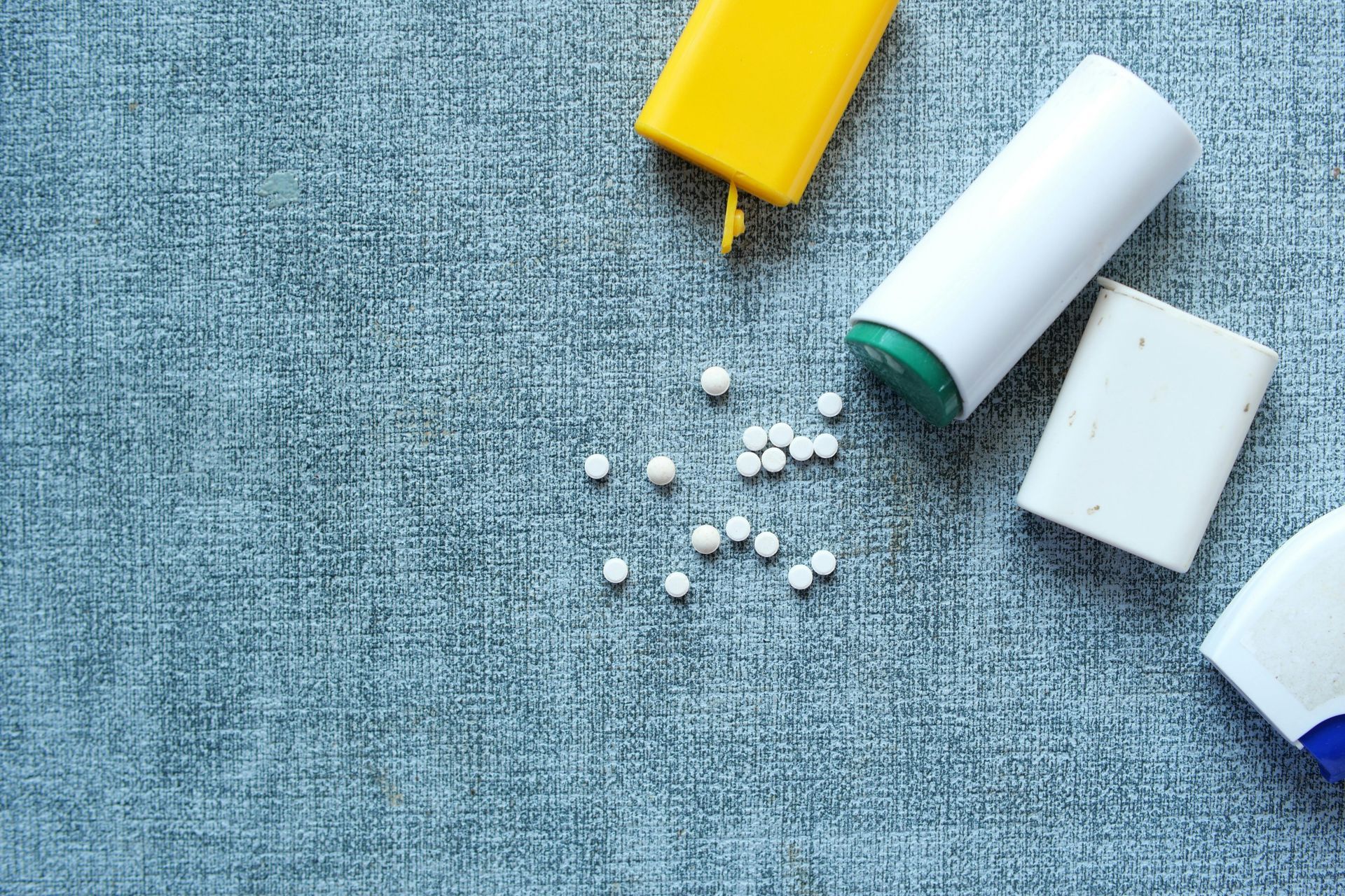 Small white pills scattered near containers, yellow, white, and blue, on a textured blue surface.