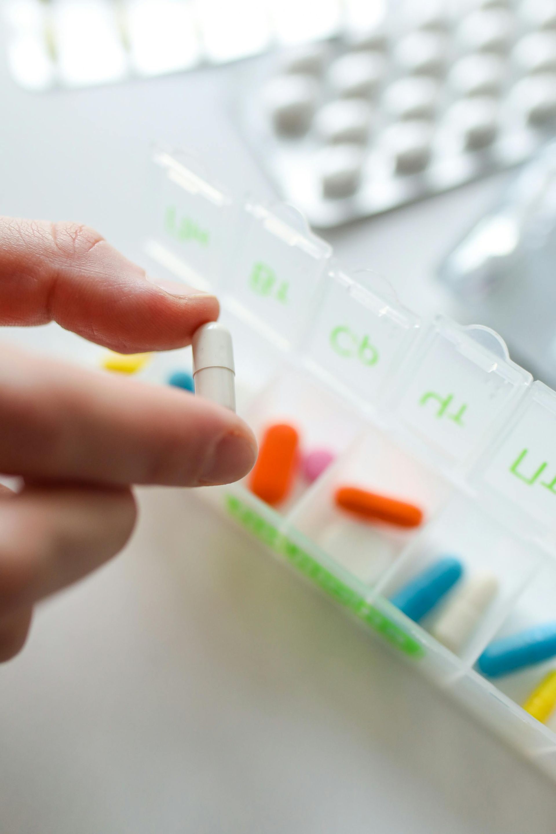Hand holding a white capsule above a pill organizer with various colored pills.