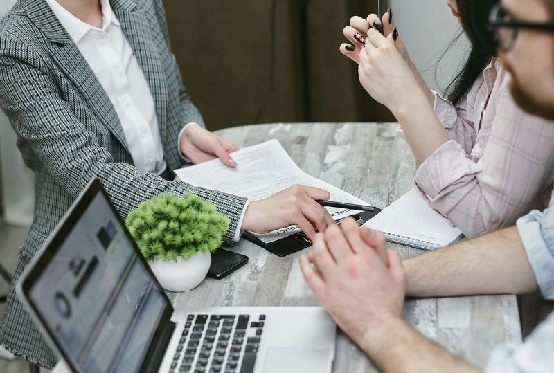 Woman in blazer reviews paperwork with couple at a table; laptop, calculator, and plant visible.