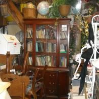 A tall, brown wooden bookshelf filled with various books stands in a cluttered room with a globe and baskets on top.
