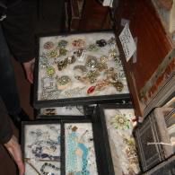 A person holds a tiered display case filled with assorted vintage jewelry, including brooches, necklaces, and rings.