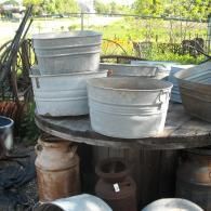 Stacked metal wash tubs and rustic milk cans arranged on a round wooden table in an outdoor setting.