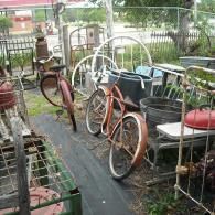 Two vintage, rusted bicycles parked in a cluttered yard filled with old metal bed frames, containers, and household goods.