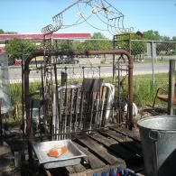 A rusted metal headboard and frame sit outdoors on a wooden pallet near a metal bucket and a shallow pan with a brick.