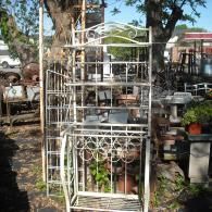 A light-colored, ornate wrought iron bakers rack standing outdoors in a salvage yard among other metal items.
