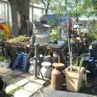 A cluttered garden area with a vintage hand water pump, assorted pots, plants, and decorative objects on stone pavers.